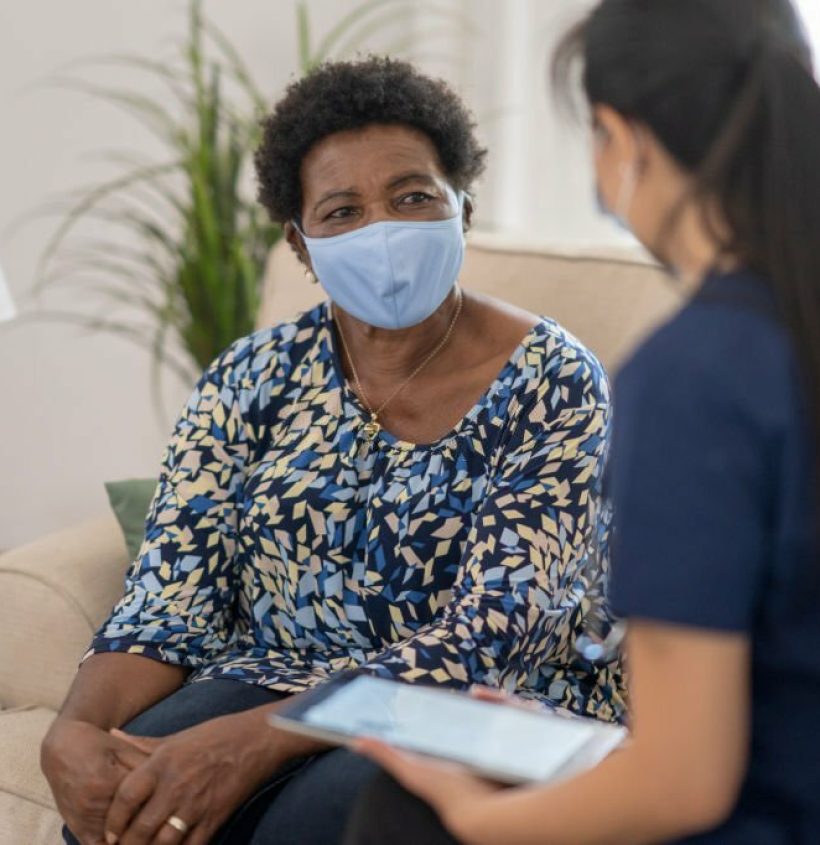 Female nurse visiting senior patient in her home during the COVID-19 pandemic, both are wearing masks to avoid the transfer of germs.